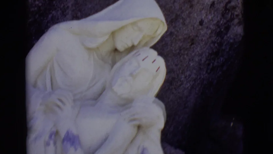 1965 8mm frame from San Luis, Colorado showing a white Pieta-like outdoor statue against a dark stone backdrop.