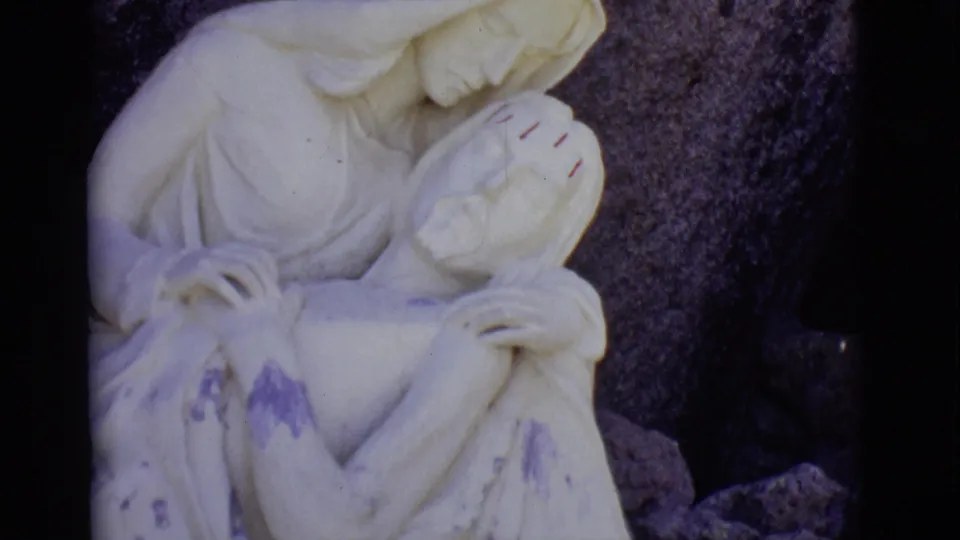 The San Luis, Colorado home-movie frame shows the Pieta-like statue more centered, with weathered marks and rocks behind it.