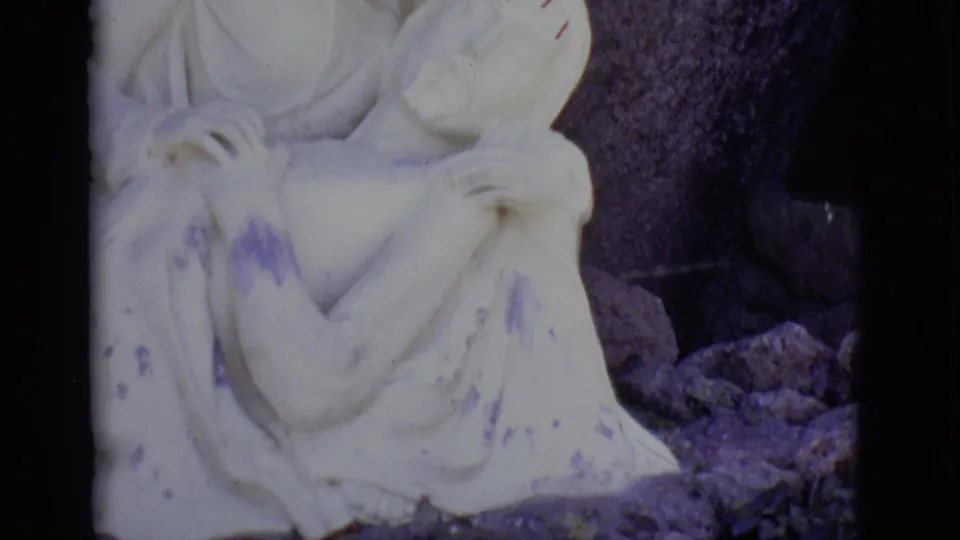 A soft 8mm close view emphasizes the statue's draped forms, purple discoloration, and surrounding stones.