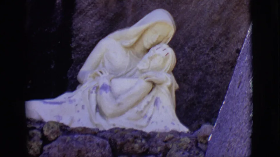 1965 8mm frame from San Luis, Colorado, showing a white Mary-and-Jesus sculpture set before a dark stone wall.