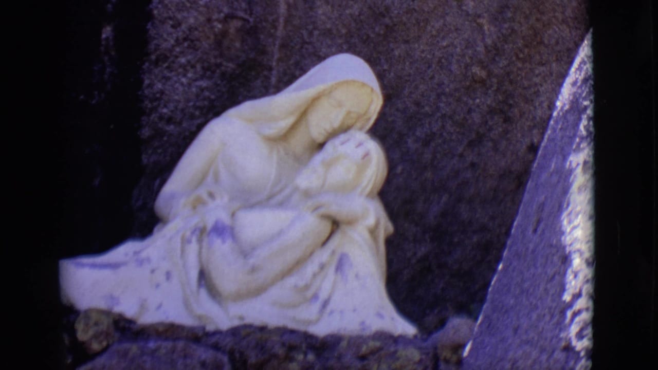 The San Luis, Colorado 8mm frame holds on the white devotional sculpture with dark rock and a slanted stone border nearby.