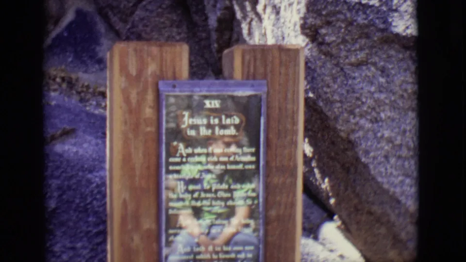 1965 San Luis, Colorado 8mm frame showing a wooden Station XIV sign reading Jesus is laid in the tomb against stonework.