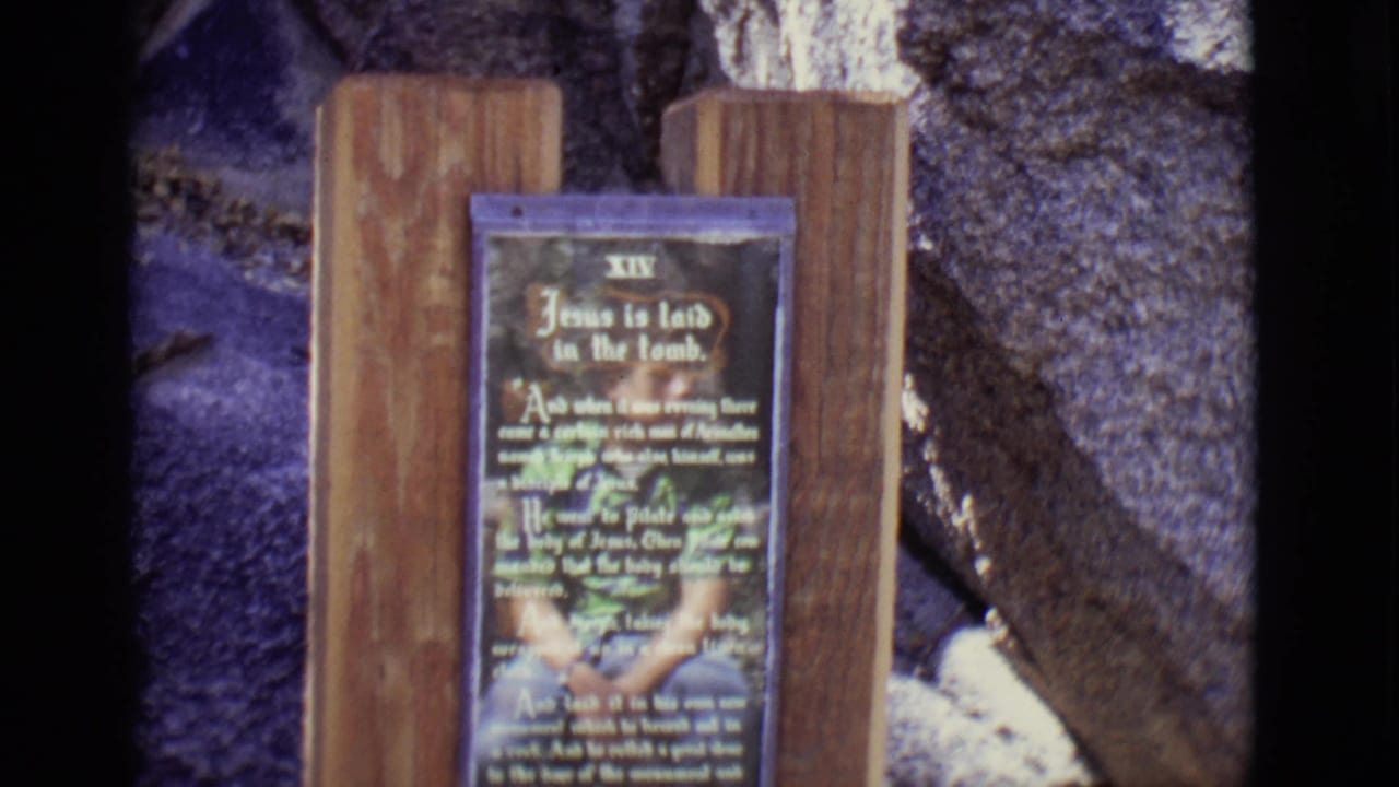 Station XIV sign framed between wood posts with stone and painted scenery behind it.