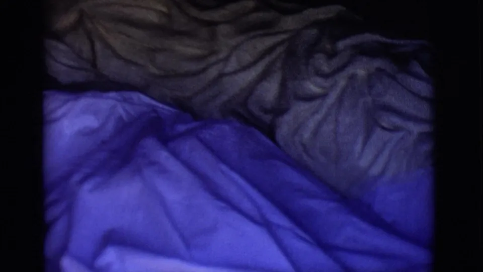 1965 San Luis, Colorado 8mm frame showing blue plastic beneath darker folded material, with no clear face visible.