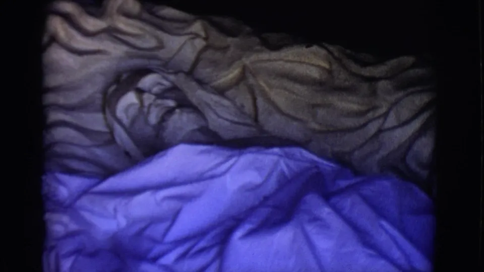 1965 8mm frame from San Luis, Colorado showing a reclining Jesus statue beneath gray and blue fabric in a dark interior-like setting.
