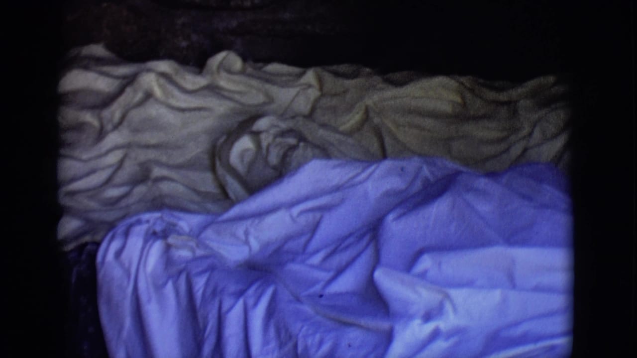 1965 San Luis, Colorado home-movie frame with blue and tan bedding seen close up, softened by grain and low exposure.