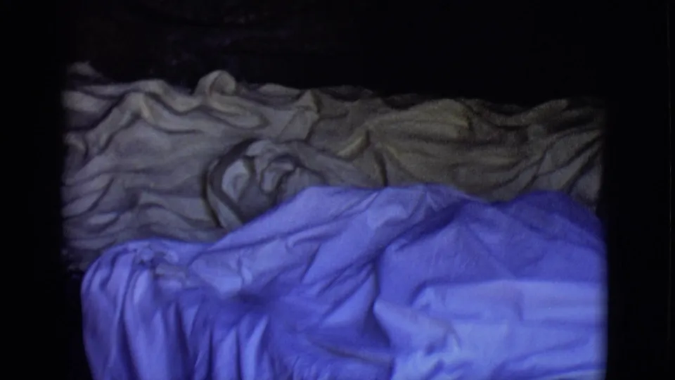 1965 San Luis, Colorado 8mm frame showing wrinkled blue and tan blankets under uneven exposure and heavy shadow.