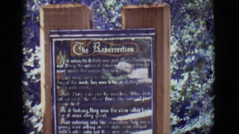 1965 8mm frame from San Luis, Colorado showing a wooden sign titled The Resurrection in a leafy outdoor setting.