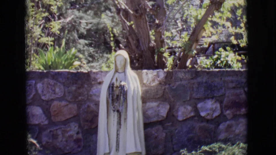 1965 San Luis, Colorado home-movie frame showing a white robed Jesus statue near a stone wall with trees behind it.