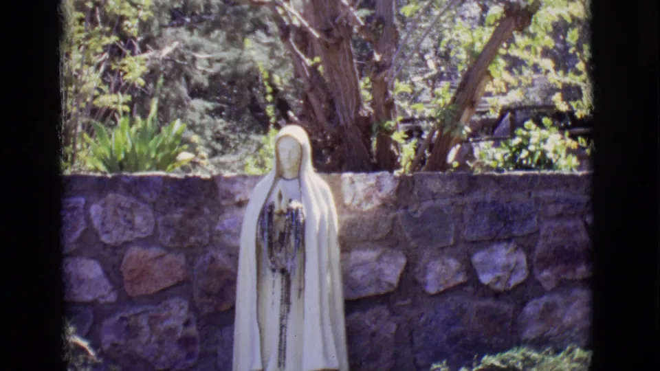 A white Jesus statue holding a cross stands in front of a stone wall and trees in this 1965 San Luis, Colorado frame.