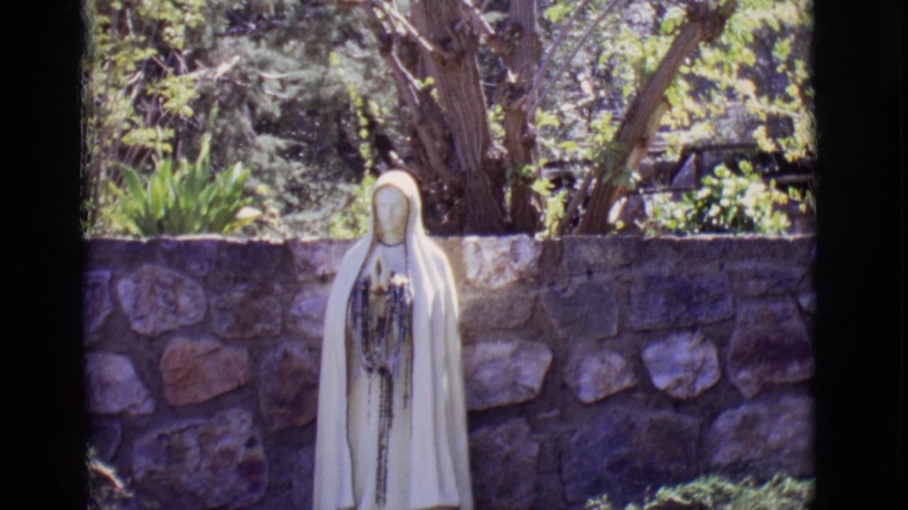 White Virgin Mary statue in a garden before a stone wall.