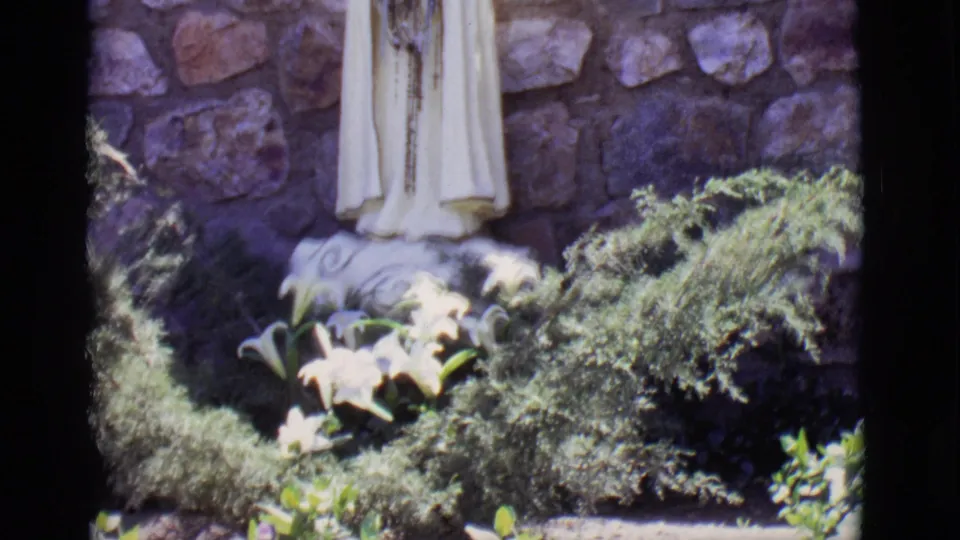 A fuller 1965 8mm view shows the white Virgin Mary statue, floral base, surrounding shrubs, and stone wall in bright outdoor light.
