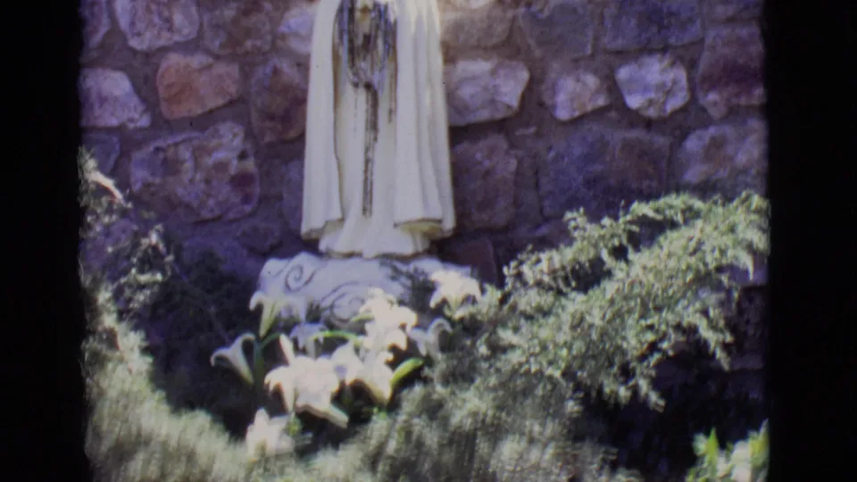 1965 San Luis, Colorado home-movie frame focusing on the lower robe of a Mary statue, white lilies, shrubs, and rough stone wall.