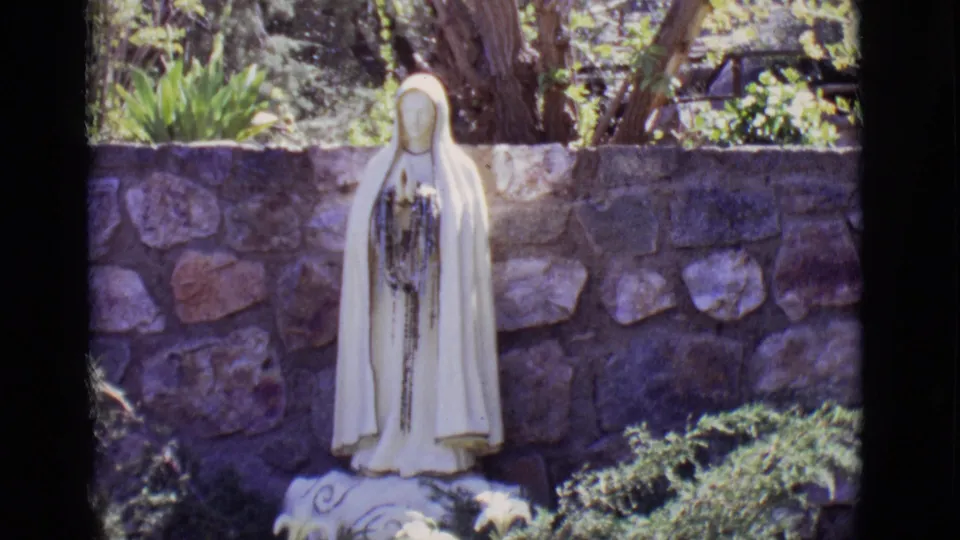 1965 San Luis, Colorado 8mm frame showing a white Mary statue in a garden, with shrubs, lilies, and a stone wall behind it.