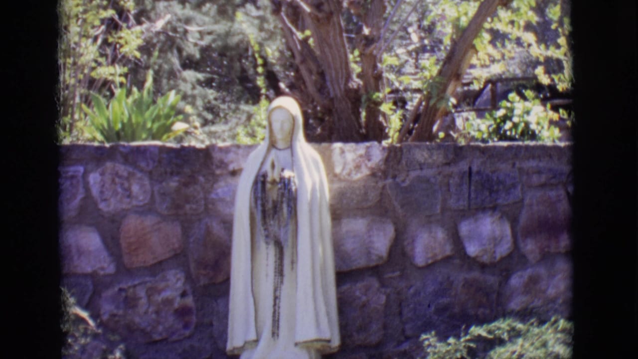 White robed Mary statue before a stone wall and garden greenery.