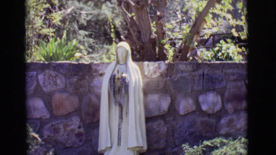 1965 8mm frame from San Luis, Colorado showing a white Mary statue with rosary-like detail in front of a stone wall and trees.