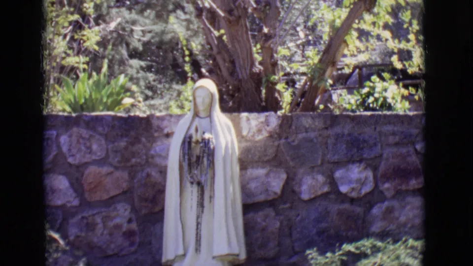 The San Luis, Colorado home-movie frame holds on the white Mary statue, with rough stonework and shaded greenery behind it.