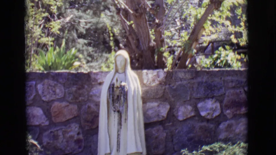 A slightly closer 1965 8mm view shows the white devotional statue partly cropped, standing before a stone wall in San Luis, Colorado.