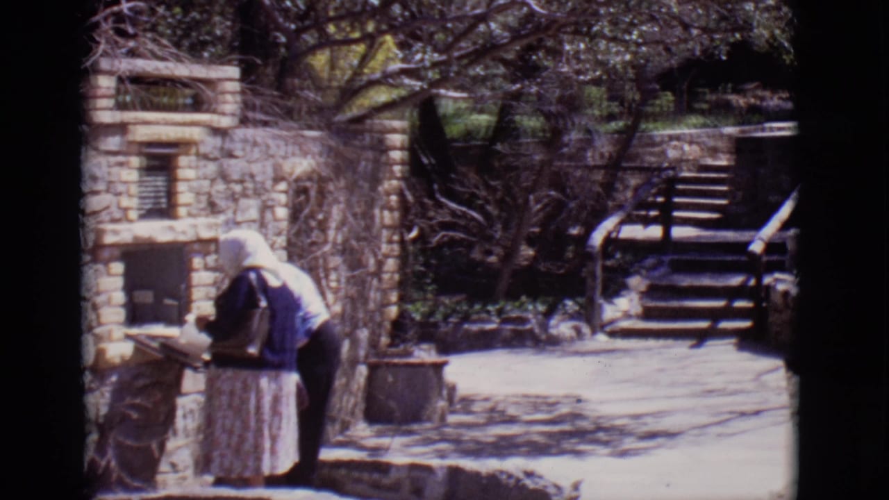 Woman in a headscarf stands beside a stone wall and small structure near outdoor steps.