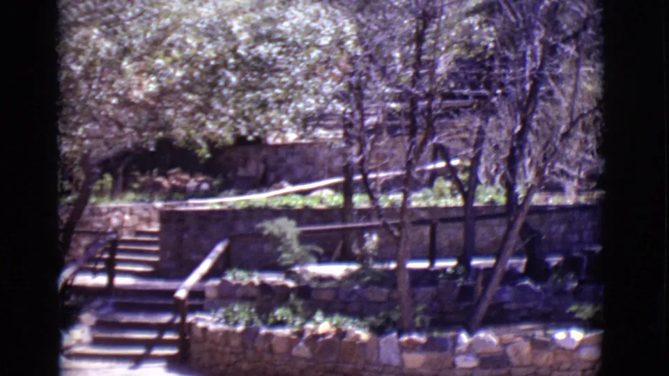 1965 San Luis, Colorado home-movie frame with stone steps, wood railings, tree trunks, low planters, and shaded stone walls.