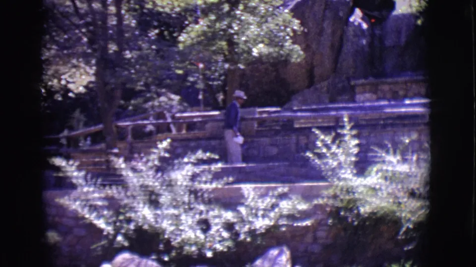 1965 San Luis, Colorado 8mm frame showing a visitor in a light hat near stone steps, railings, foliage, and a large rock formation.