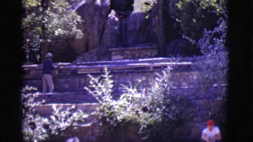 1965 San Luis, Colorado 8mm frame showing a hat-wearing visitor beside stone steps, overgrown plants, railings, and a rocky backdrop.