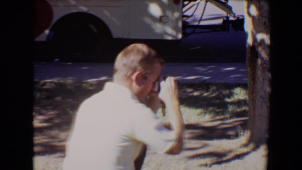 In 1967 Evansville, Indiana home-movie footage, an adult man in a white shirt crouches in dappled shade and appears to take a photograph with a small camera.
