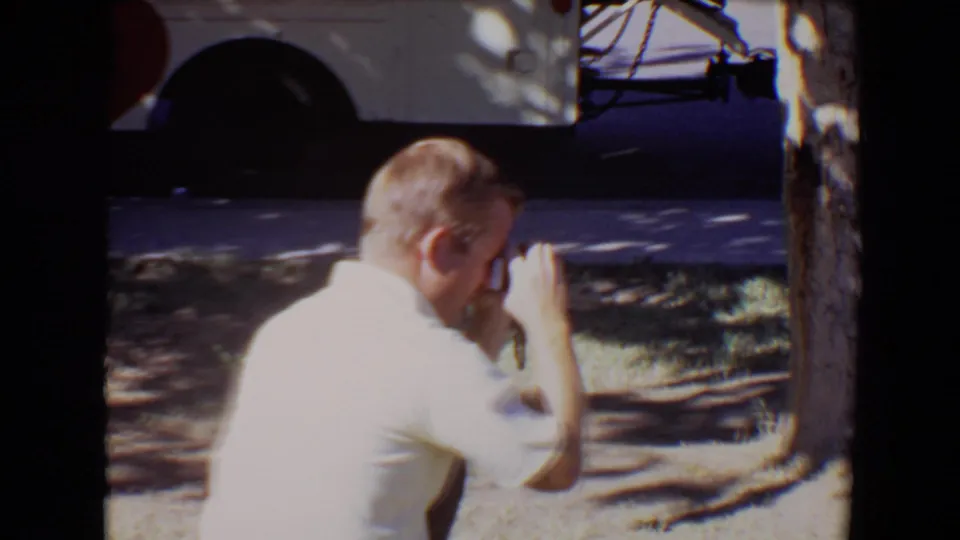 In 1967 Evansville, Indiana home-movie footage, an adult man in a white shirt crouches in dappled shade and appears to take a photograph with a small camera.