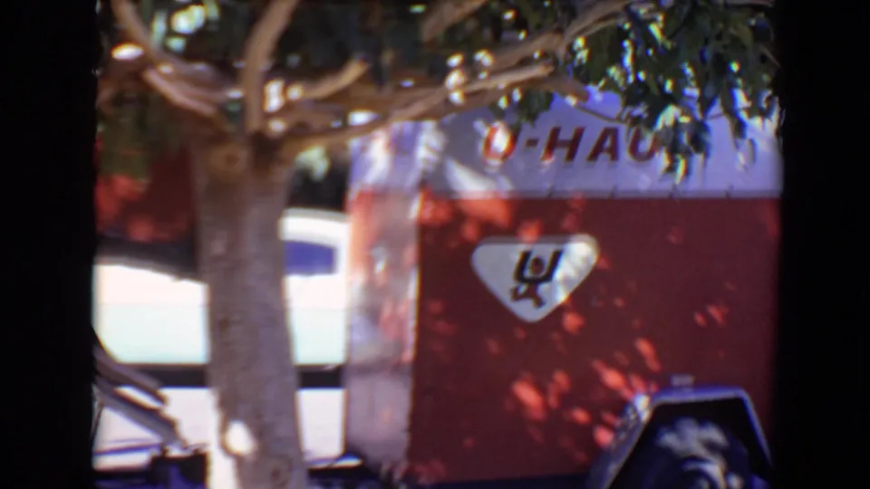 A red-and-white U-Haul trailer stands behind tree trunks and leaves, with partial lettering and logo visible in the 1967 Evansville home-movie frame.