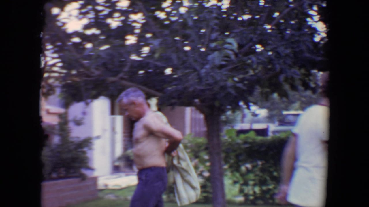 Shirtless man walks past trees carrying a striped shirt in a residential yard.