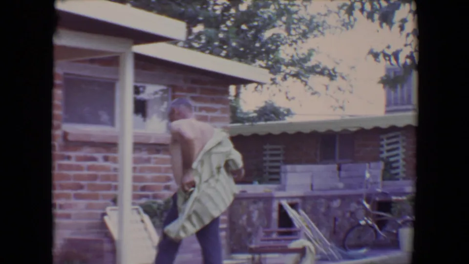 The man begins putting on the striped shirt beside the brick house, with patio furniture and a bicycle visible in the background.