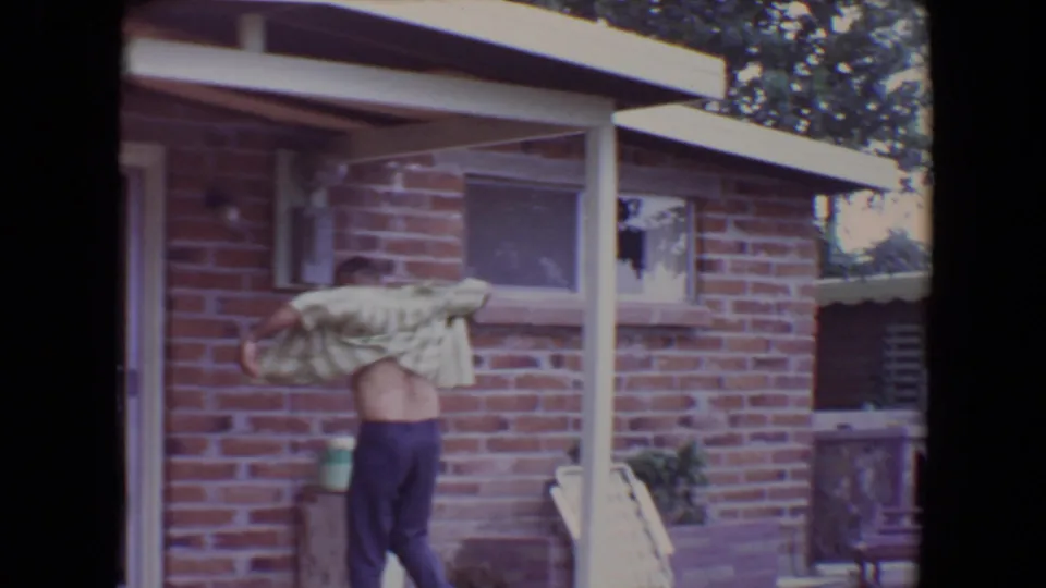 The man pulls the striped shirt over his head in a shaded residential yard, framed by porch posts and a brick wall.
