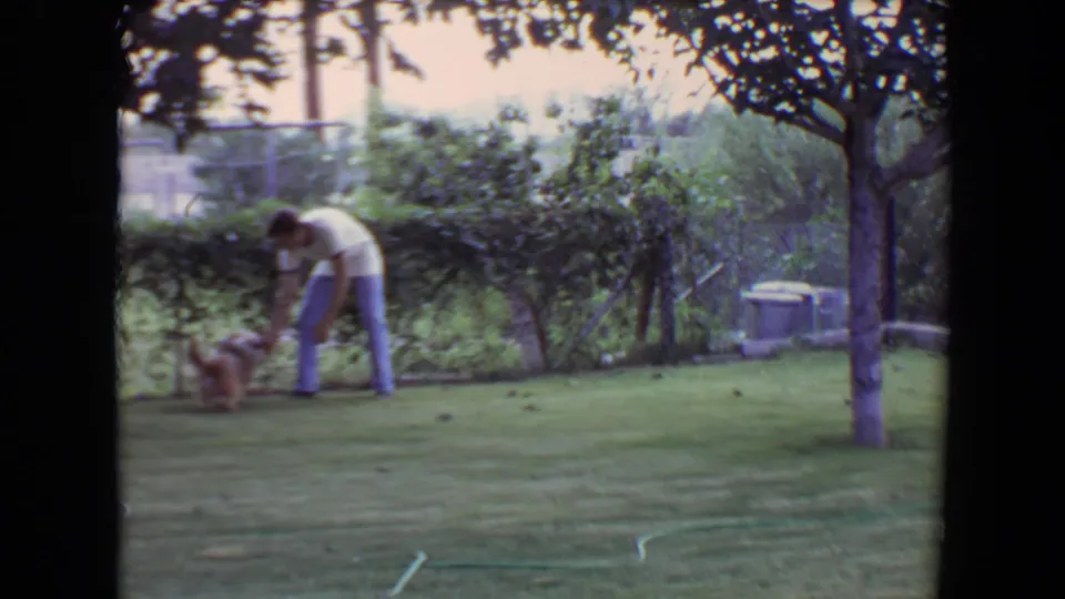 A man in a white shirt and blue jeans bends toward a small dog in a 1961 Flagstaff backyard scene.