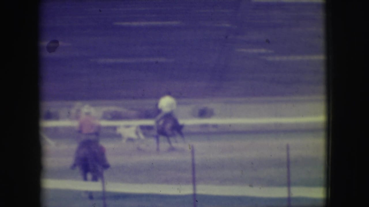Two mounted riders on a dirt track with fencing in the foreground.