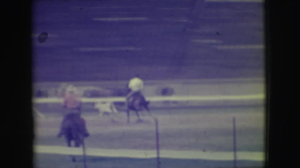 Two riders appear on a dirt track in Denver, Colorado, with fencing and distant spectators visible in the soft 8mm frame.