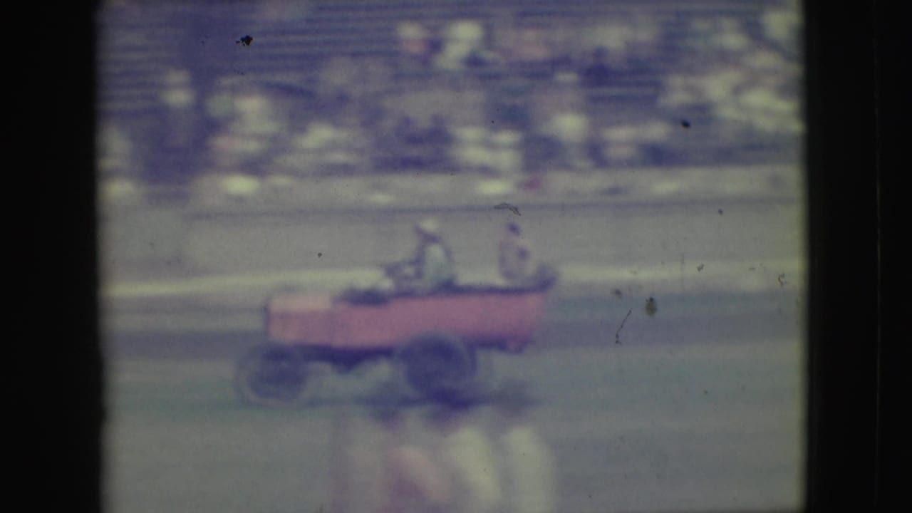 A red open vehicle with seated passengers crosses the track area in the 1949 Denver home-movie sequence.