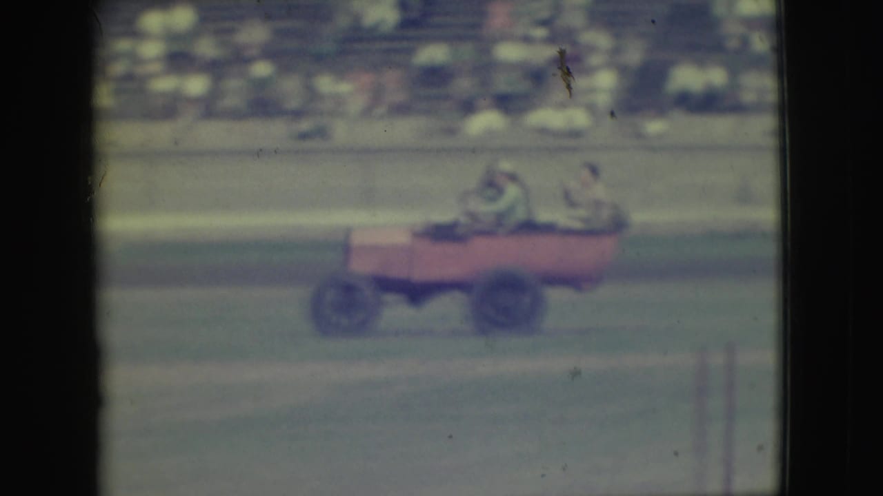 The red open vehicle continues across the Denver dirt track, with passengers seated inside and spectators blurred in the background.