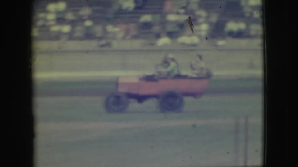 The red open vehicle continues across the Denver dirt track, with passengers seated inside and spectators blurred in the background.