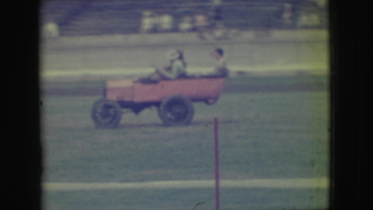 Small pink open car carries riders across a grassy field with bleachers behind it.