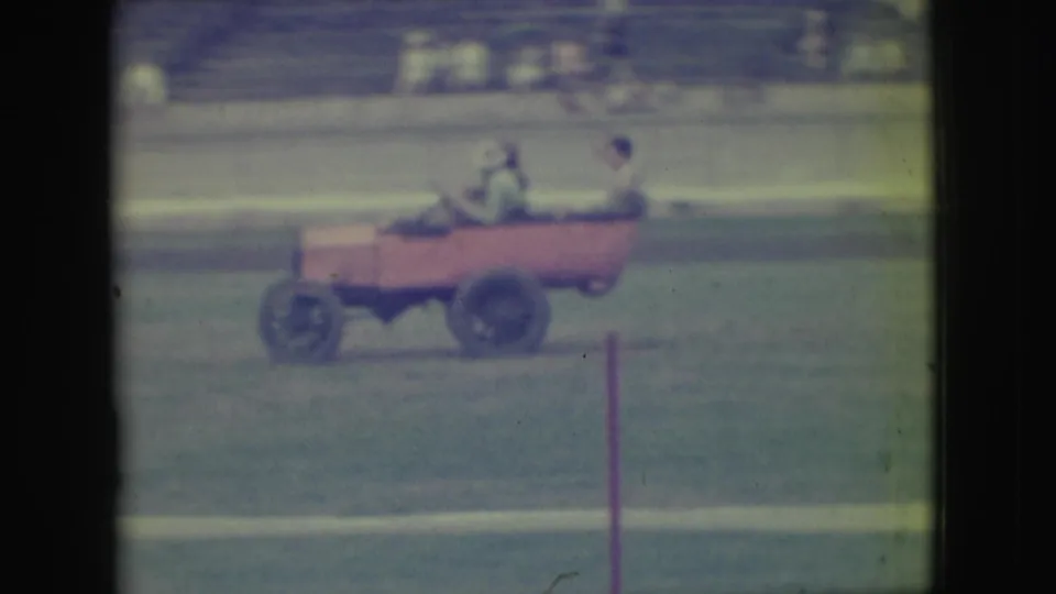 In the supplied 1949 Denver clip, a small pink open car moves across a grassy field while adult riders sit inside and bleachers blur in the background.