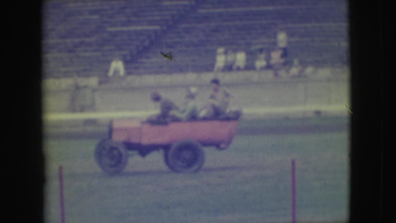 Pink open car bounces along the field with riders leaning inside and spectators in the background.