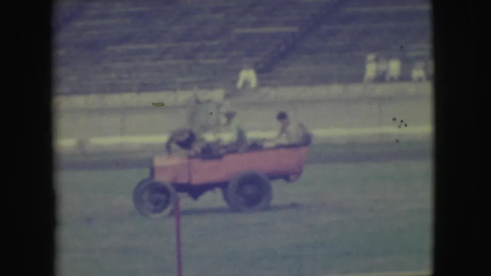 Three adult riders appear jostled in the small pink car on the field, with one figure leaning forward and blurred bleachers behind them.