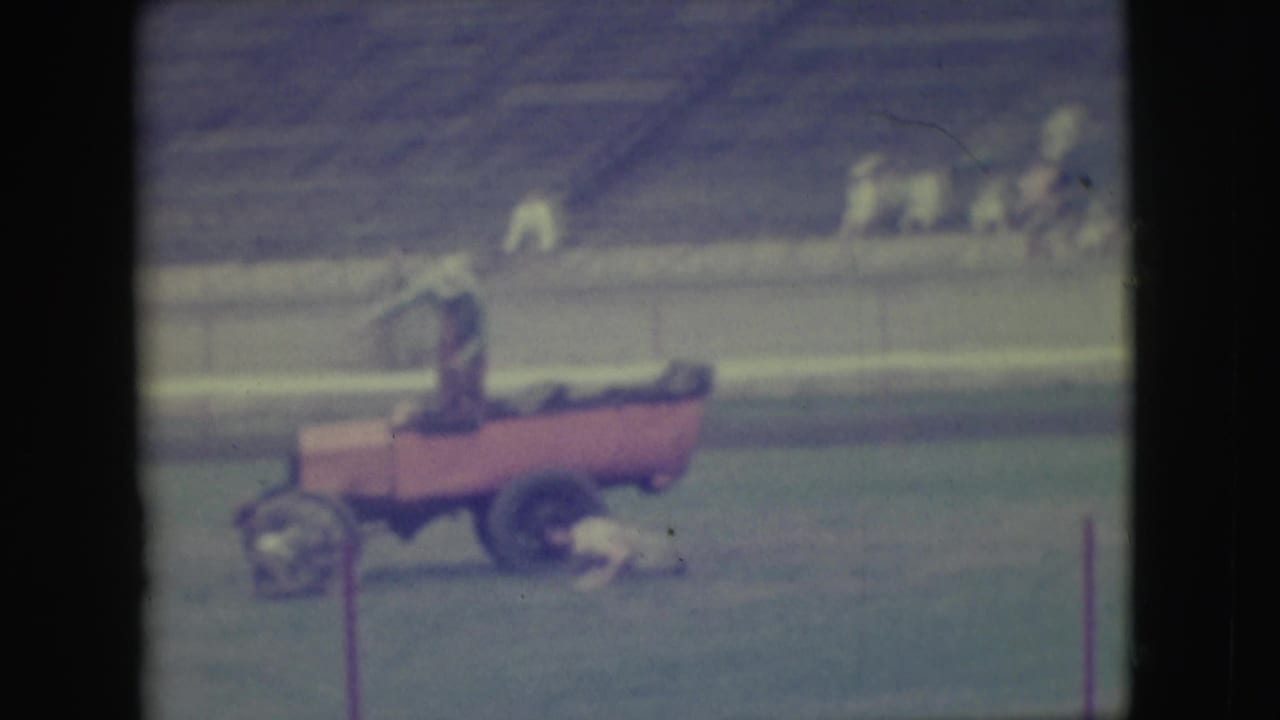 Person rides a small red cart on a baseball field with empty bleachers behind.