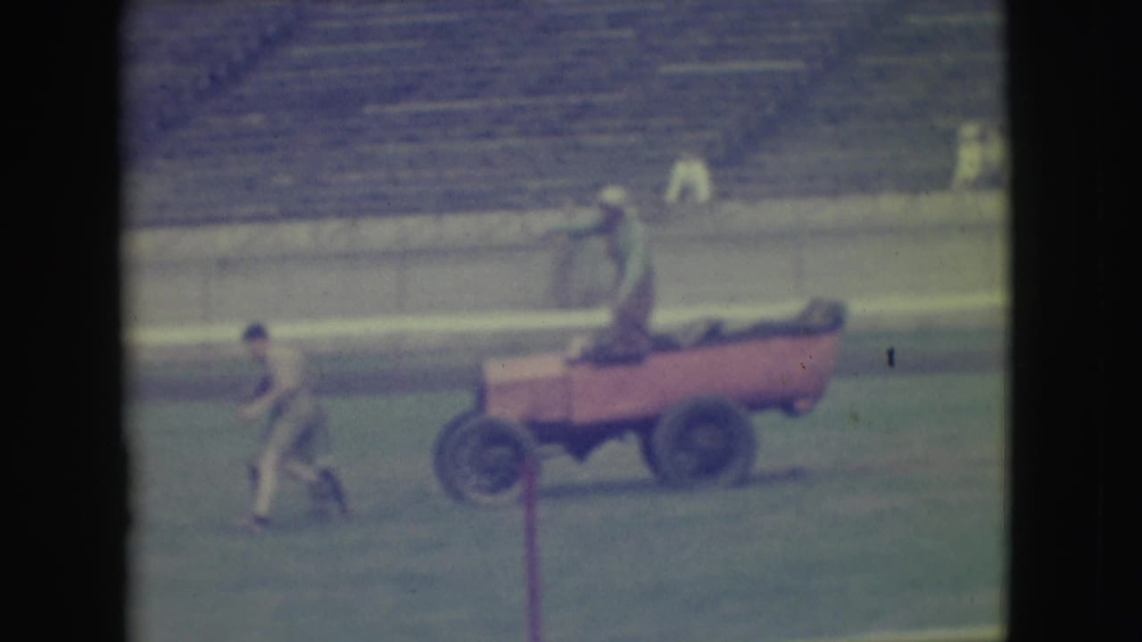 A distant 1949 Denver frame shows a person on a red cart and another person walking across the baseball field near empty bleachers.