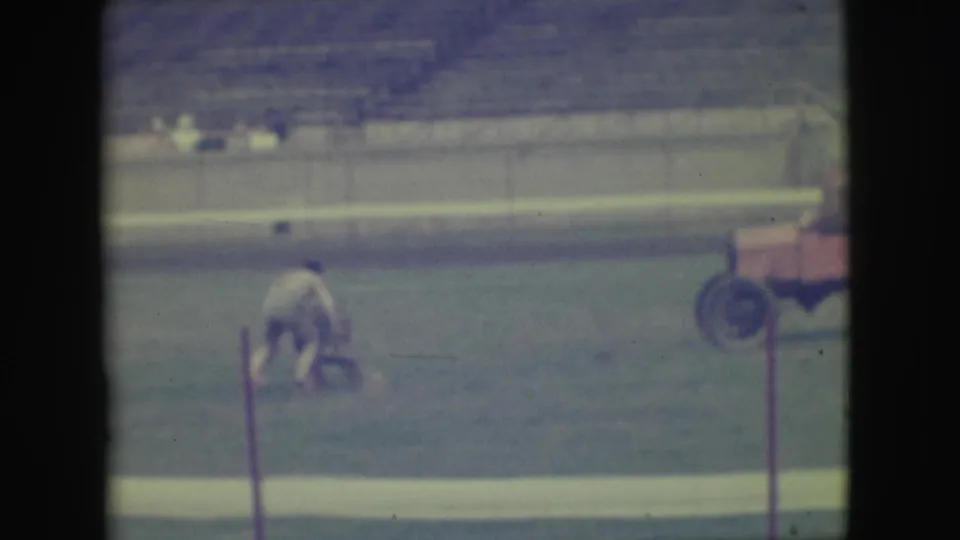 A soft 8mm frame from the Denver field shows one person bending on the grass as the red cart exits or sits partly out of frame.