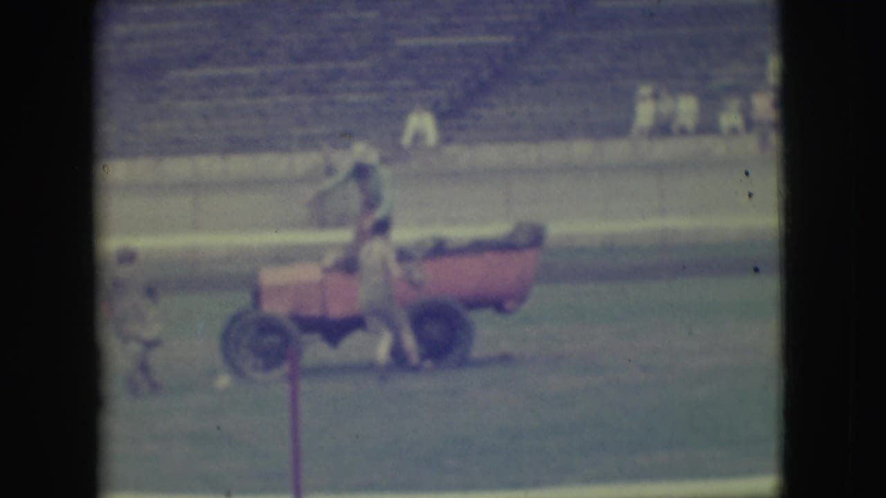 In metadata-supported 1949 Denver footage, people gather around a red open car on a stadium field with bleachers behind them.