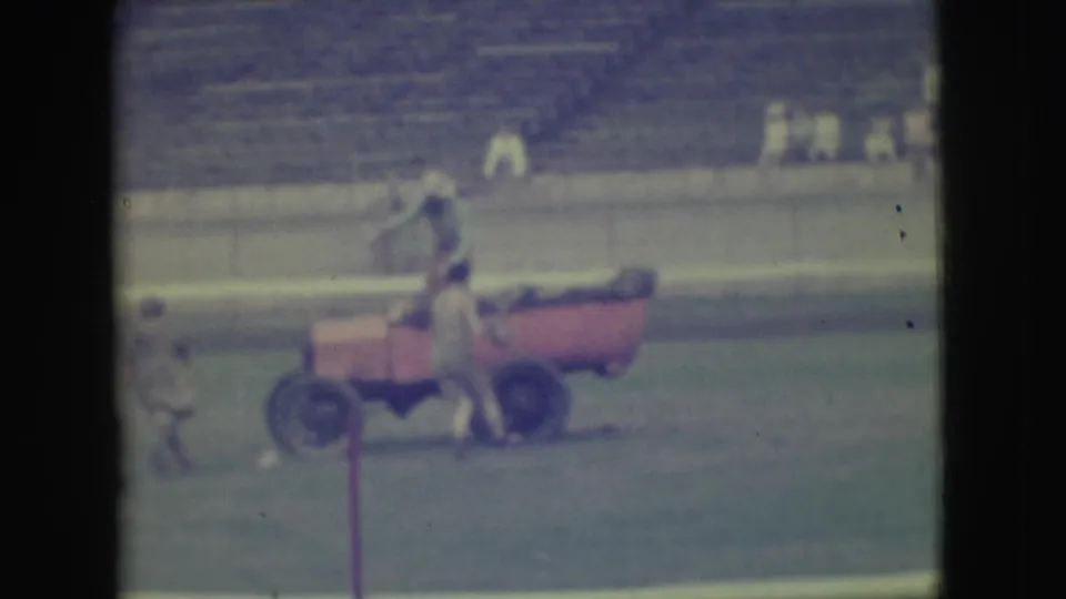 In metadata-supported 1949 Denver footage, people gather around a red open car on a stadium field with bleachers behind them.