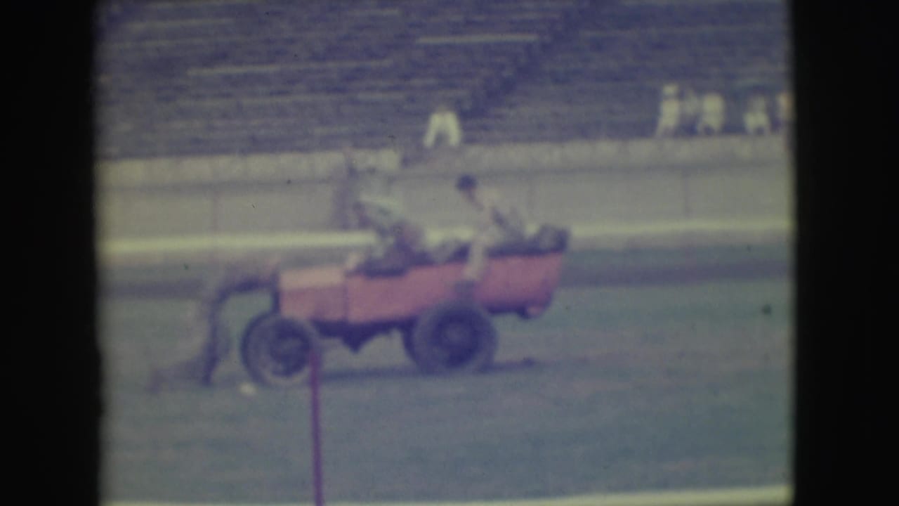 A red open car carries people across a grassy field in front of stadium seats.