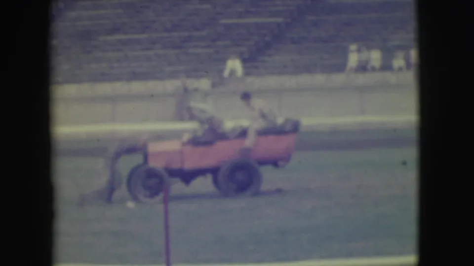 The red open car moves across the Denver stadium field with people aboard, filmed from a distant 8mm home-movie viewpoint.