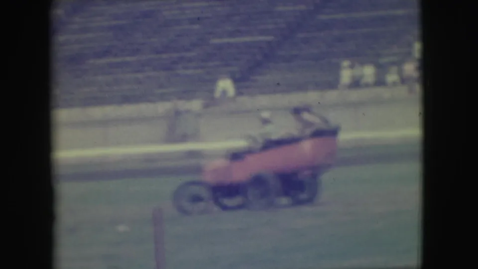 During the stunt, the red open car appears to lift its front end on the grassy stadium field as people remain close to or inside it.